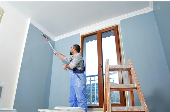 Professional painter in blue coveralls applying blue paint on a wall with a paint roller inside a bright room with wooden door frames.