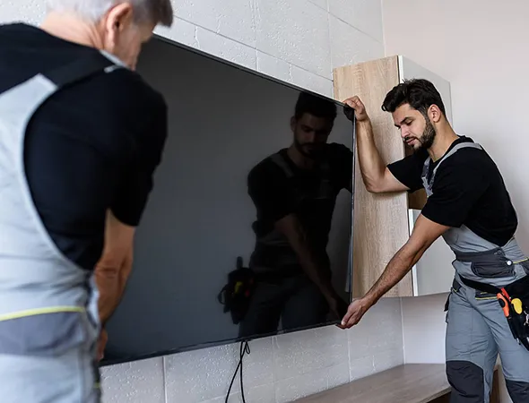 Two professionals mounting a large flat-screen TV on a living room wall, one holding and aligning the TV, the other securing it to the mount.