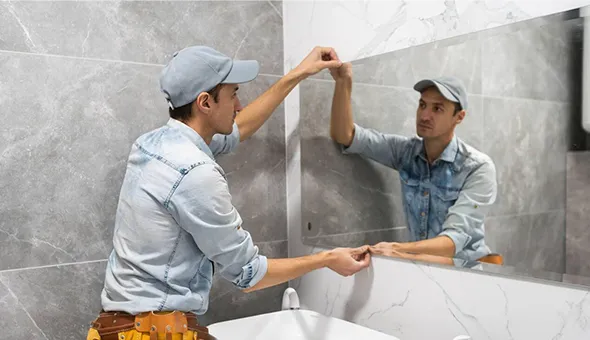 Professional installer wearing a cap and tool belt mounting a large mirror on a tiled wall in a commercial or bathroom setting.