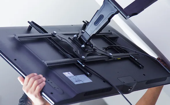 Technician holding a flat-screen TV from behind, showing the underside mounting bracket being secured to the TV.