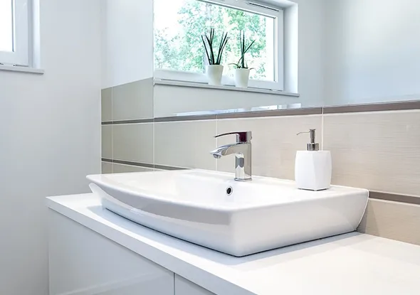 Bright residential bathroom sink setup with a white rectangular vessel sink, chrome faucet, soap dispenser, and a small plant by a window letting in natural light.
